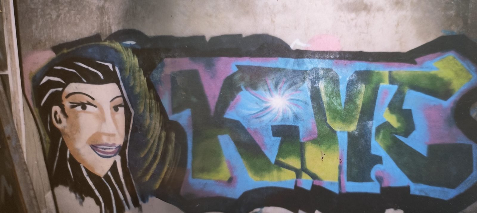 Fresque sur un mur : un visage de femme aux longs cheveux noirs, à côté d'un grand lettrage graffiti vert et bleu.