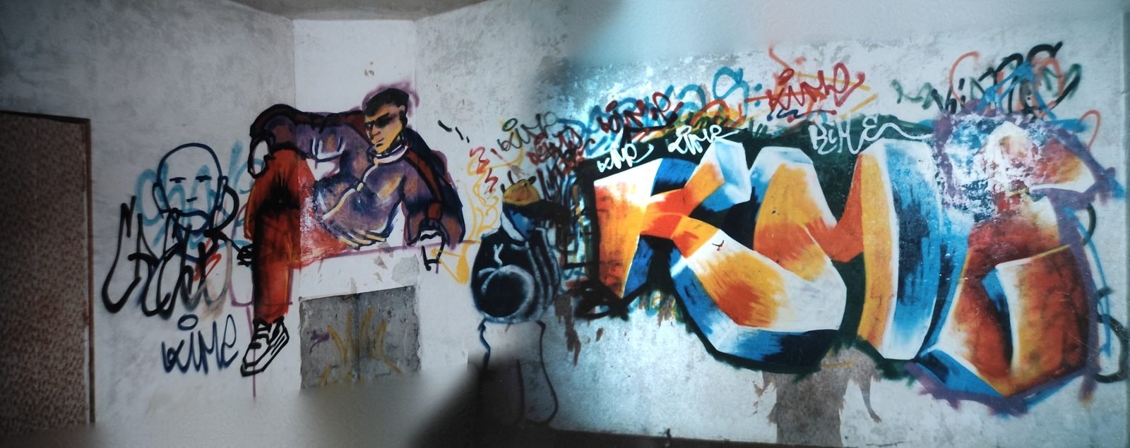 Fresque sur un long mur : un personnage en lunettes noires et blouson, puis un grand lettrage graffiti orange et bleu.