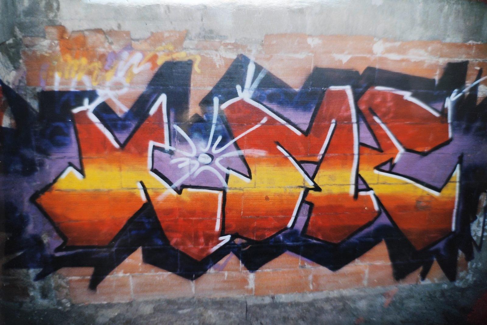 Lettrage graffiti en dégradé orange et jaune, contour violet et reflets blancs, peint sur un mur de briques.
