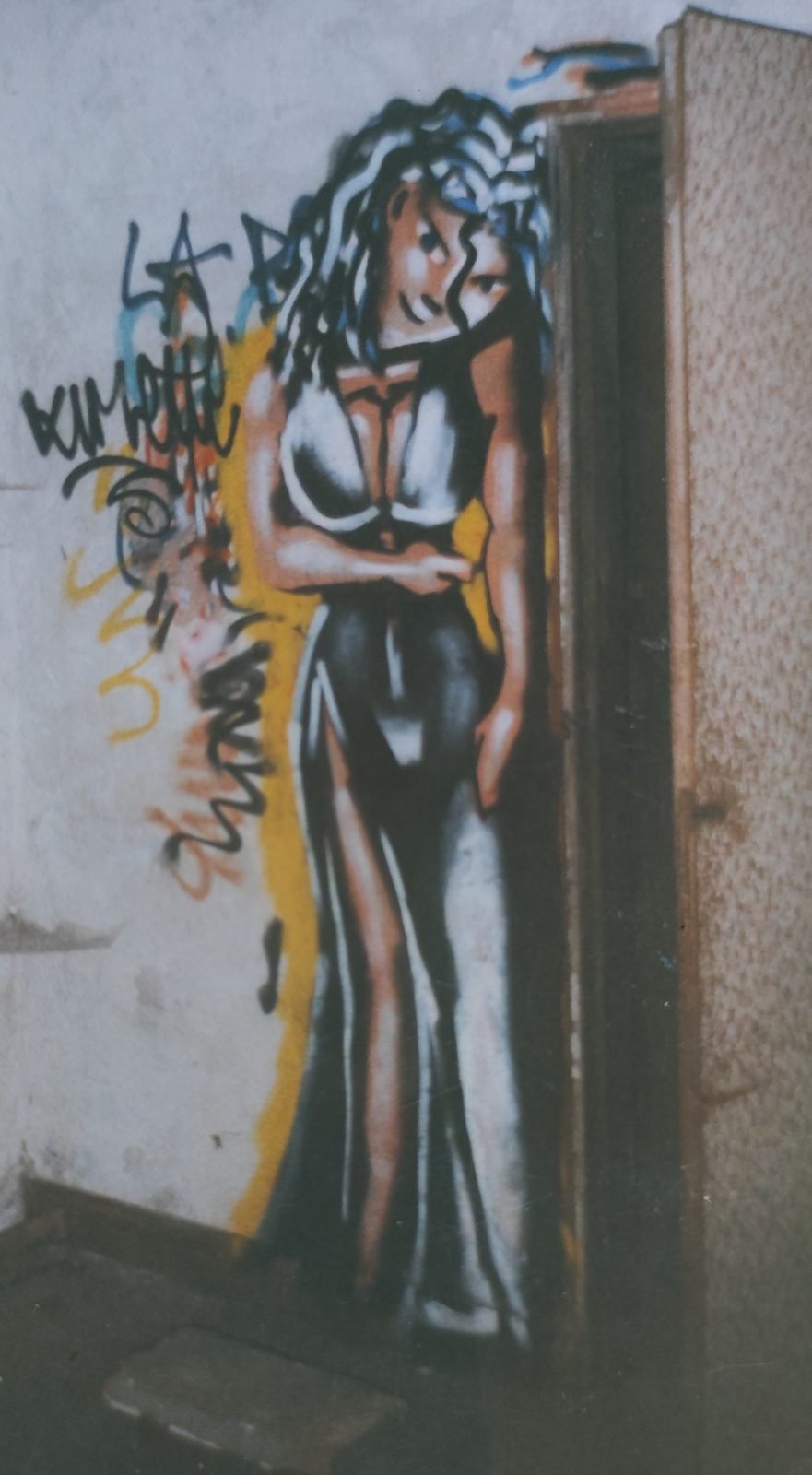 Graffiti d'une femme en longue robe noire décolletée, adossée à l'encadrement d'une porte, sur un mur blanc.