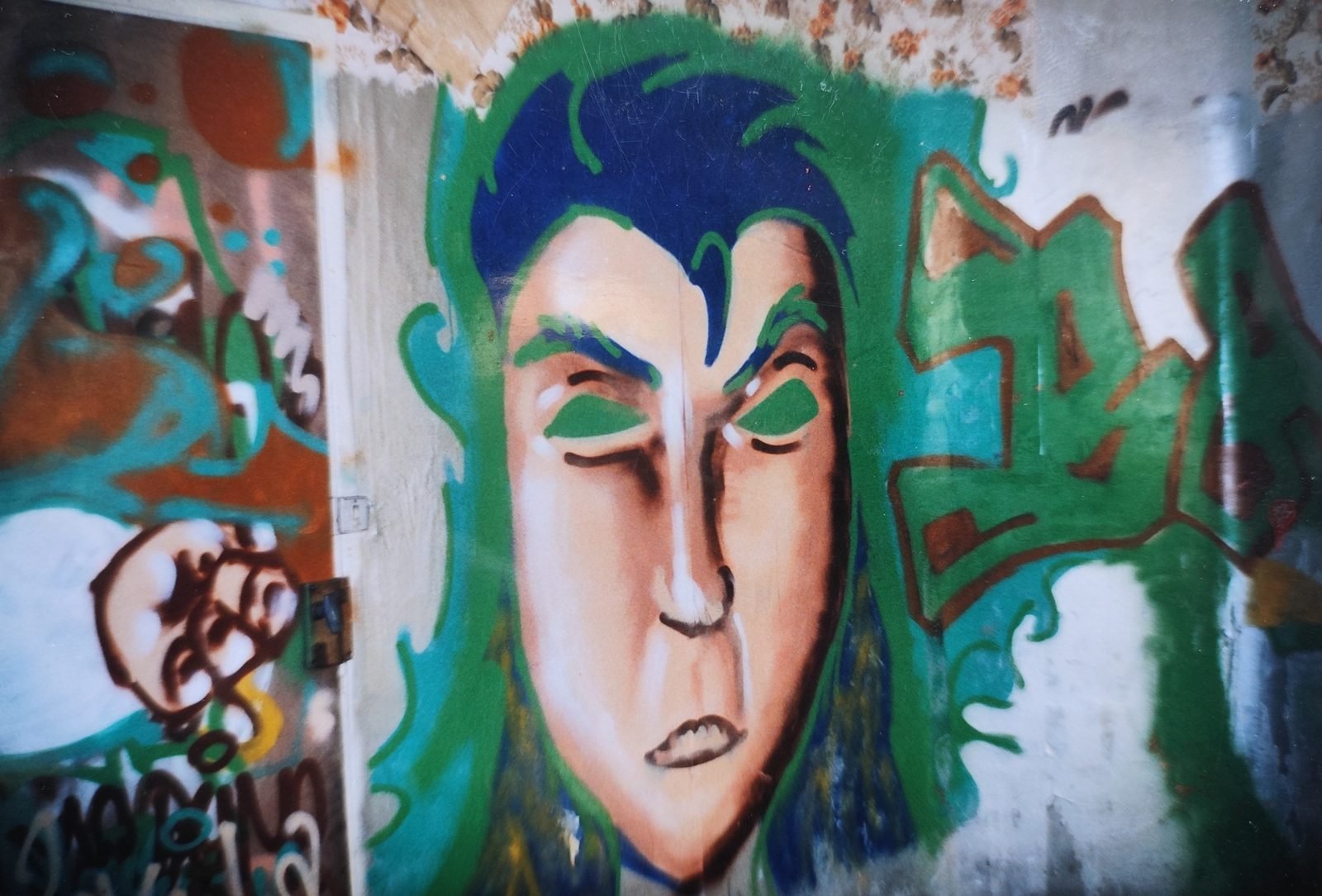 Graffiti d'un grand visage aux cheveux bleus et aux paupières vertes, entre des lettrages graffiti, sur un mur.
